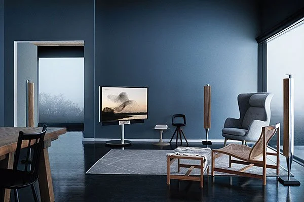 Modern living room with blue walls, a large window, a flat-screen TV, a high-back armchair, a small ottoman, two tall speakers, a side table, and a gray area rug.