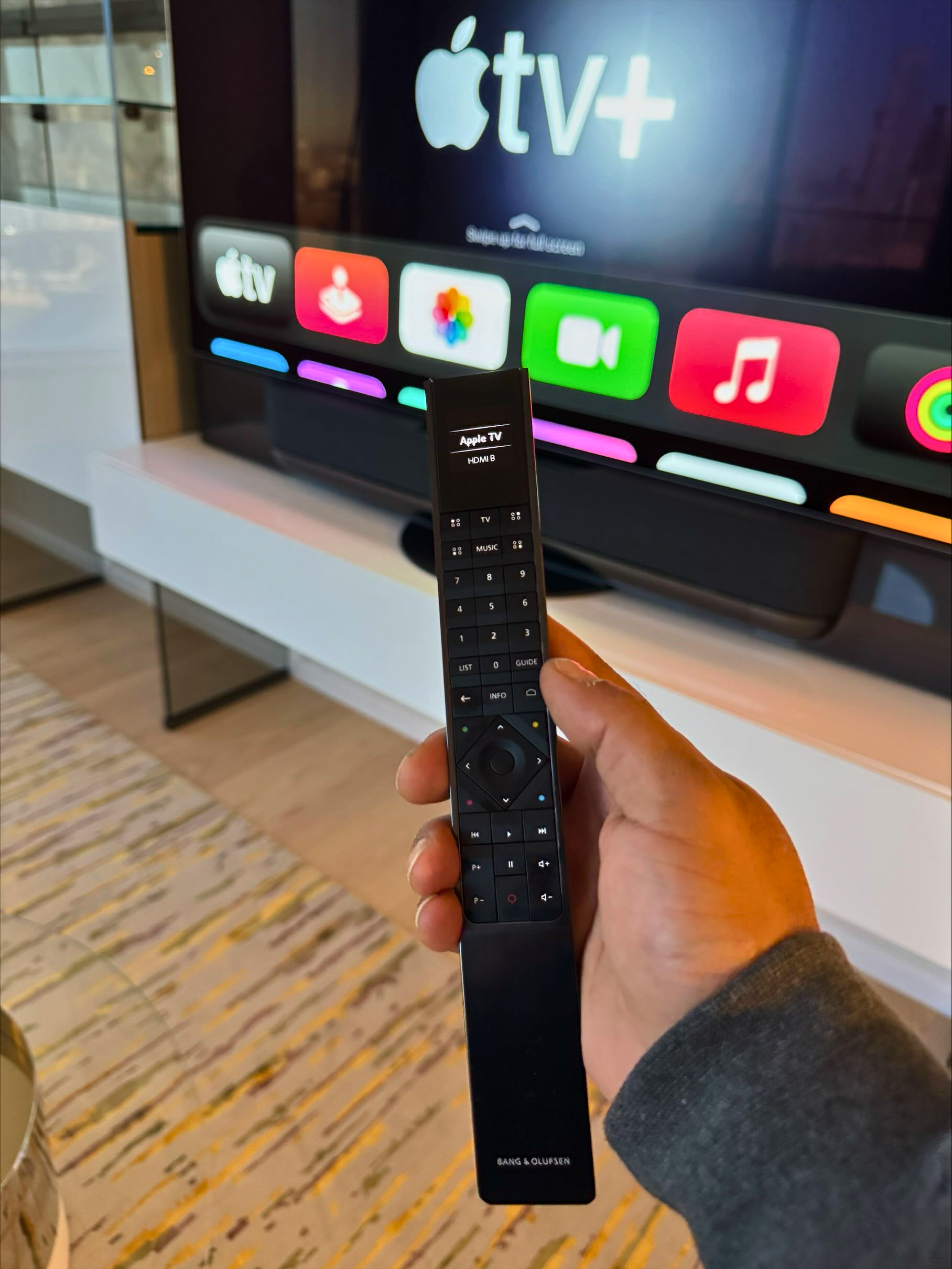 Hand holding a black Apple TV remote in front of a large screen displaying the Apple TV+ interface and app icons.