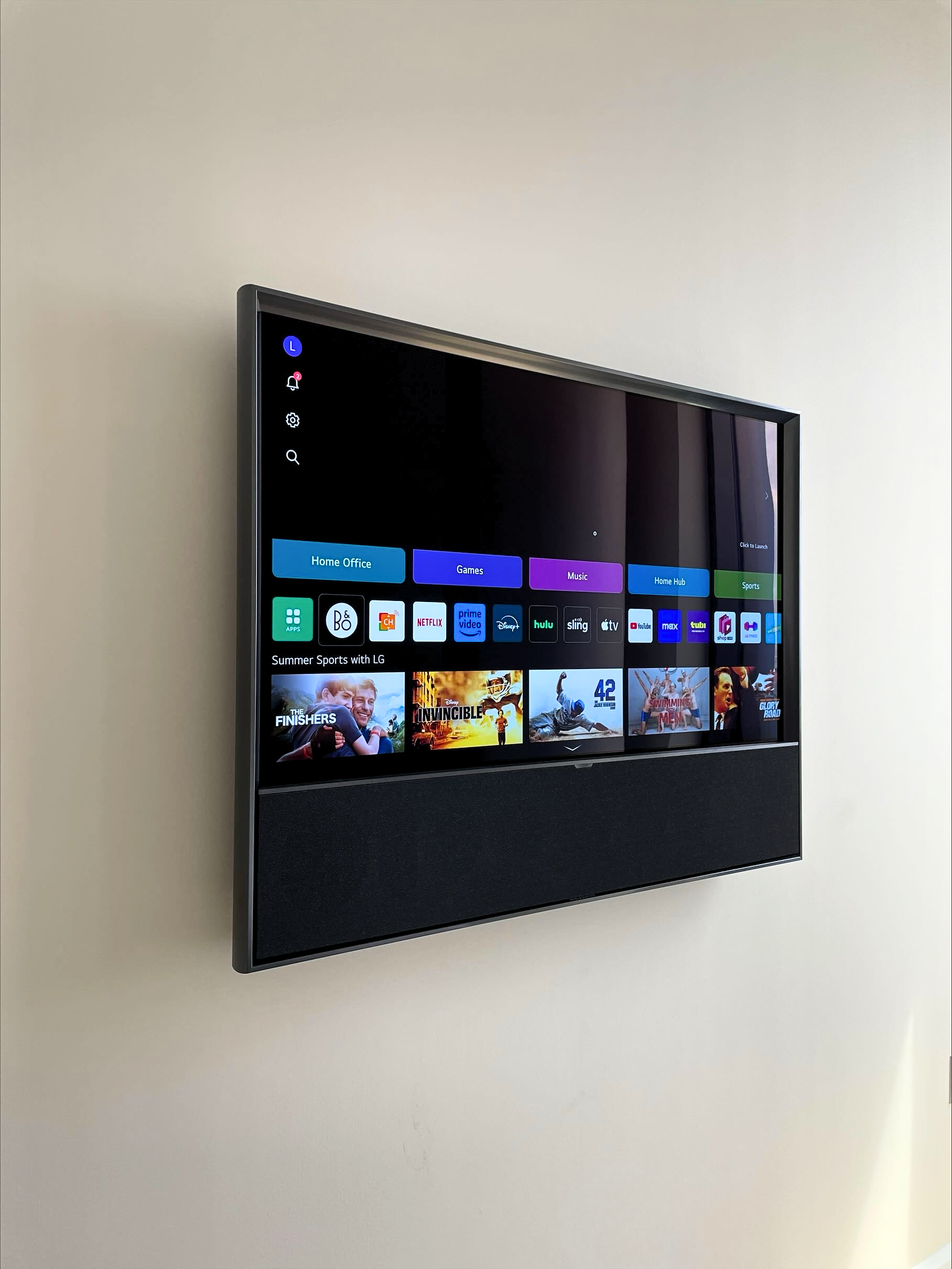 Wall-mounted LG smart television displaying a menu with categories like Home Office, Games, Music, Home Hub, and Sports, along with app icons and a selection of summer sports movies.