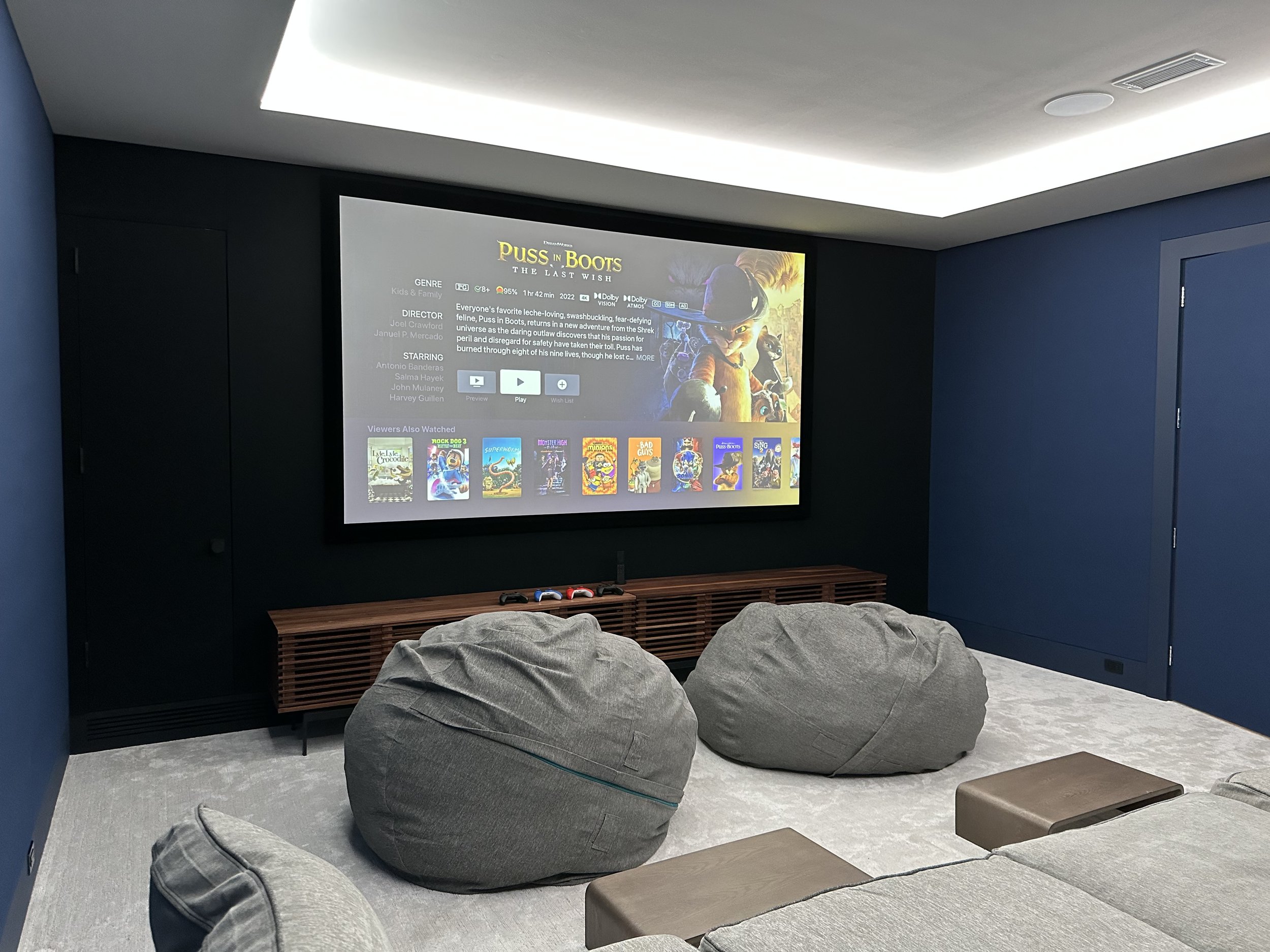 Modern home theater room with a large screen displaying a movie selection, grey bean bag chairs, and a low wooden entertainment console.