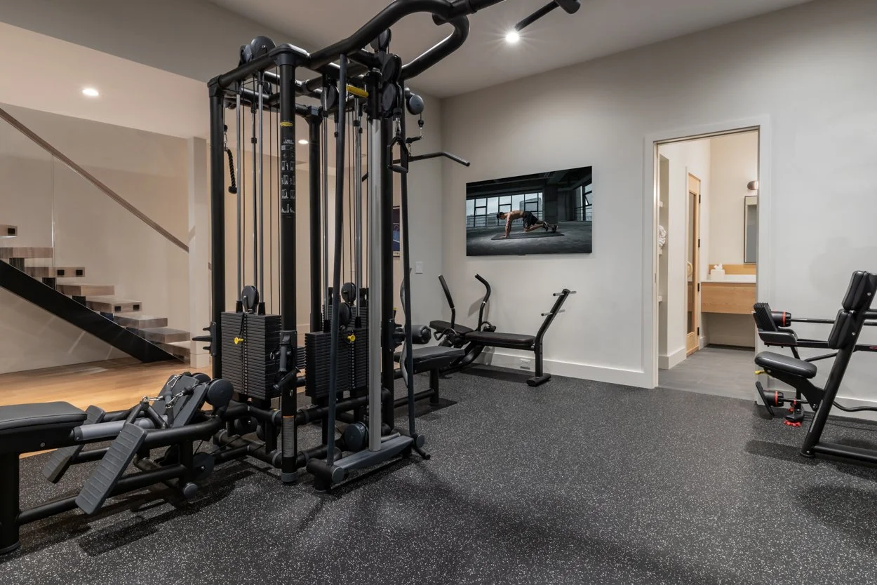 Indoor gym with various exercise equipment including a multi-station weight machine, a rowing machine, and a stationary bike. There is a flat-screen TV on the wall and an open doorway leading to a bathroom area.