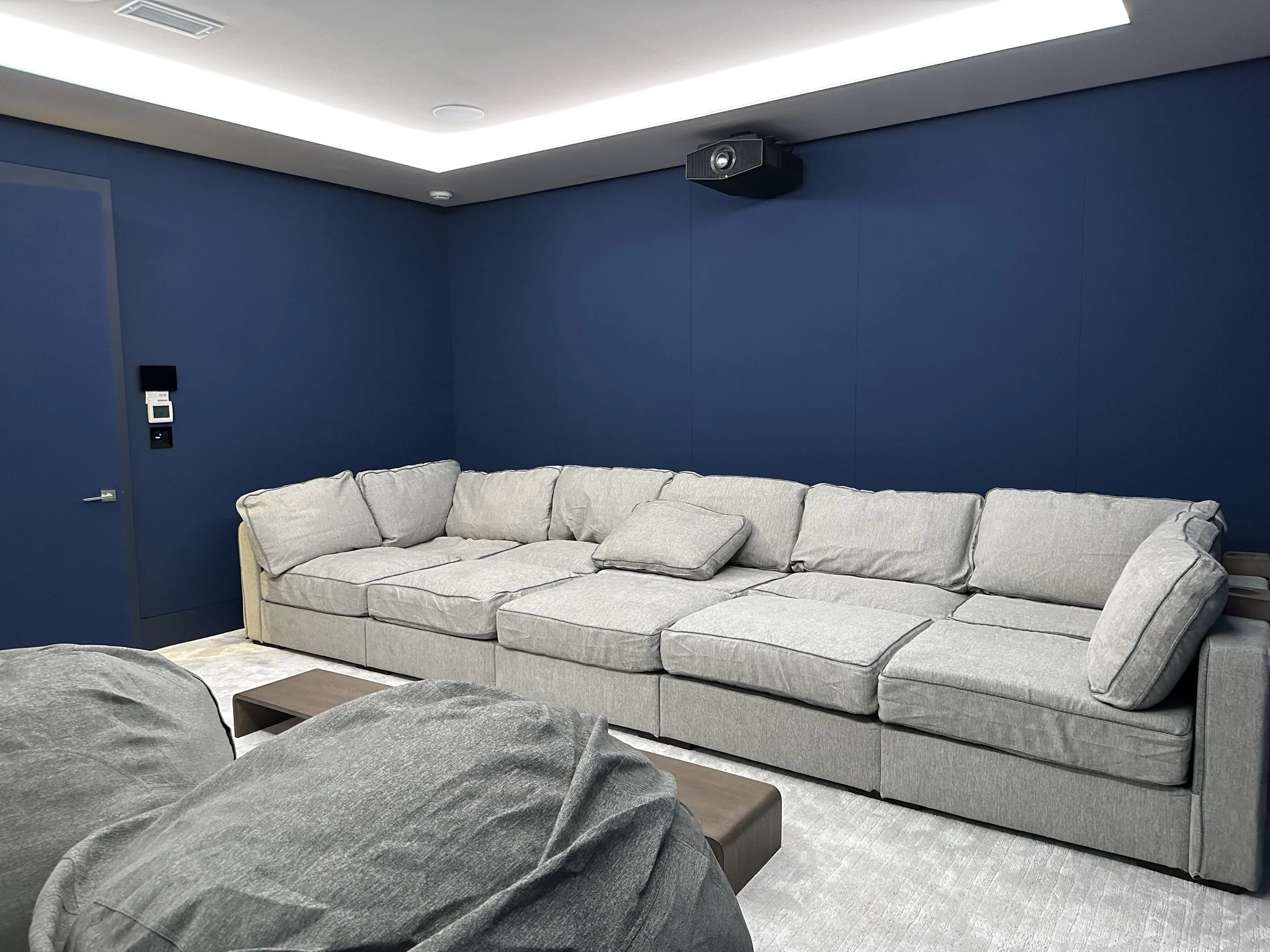 A large beige sectional sofa with multiple cushions in a room with dark blue walls, a ceiling-mounted projector, and a small dark brown coffee table.