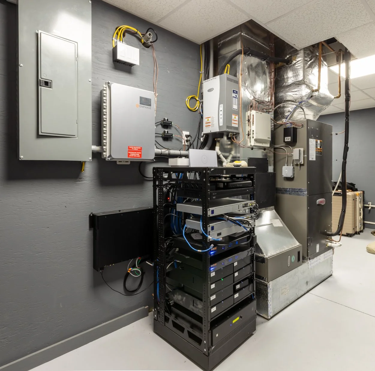 Electrical and networking equipment on a gray wall, including circuit breaker panels, servers, and various control boxes.