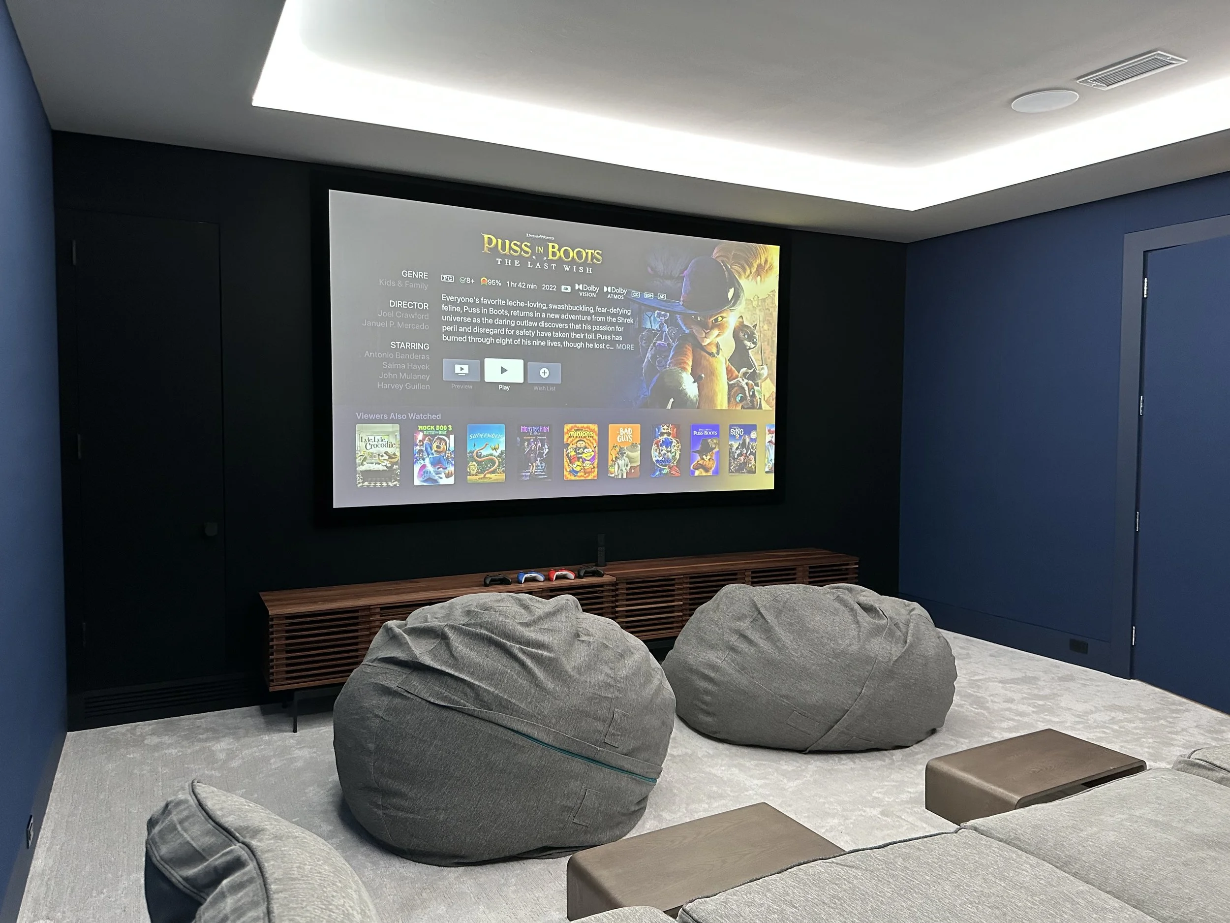 Home theater room with a large projection screen displaying the movie 'Puss in Boots: The Last Wish', surrounded by gray bean bag chairs and a low wooden media console underneath the screen.