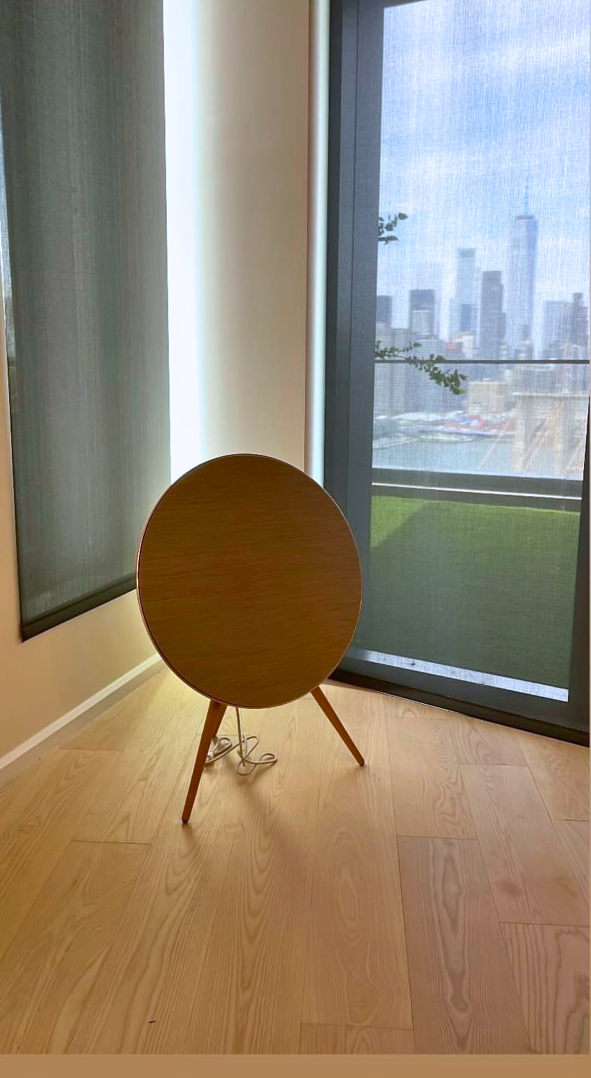 A small, round wooden table with three legs, placed near a glass door with a cityscape view in the background.
