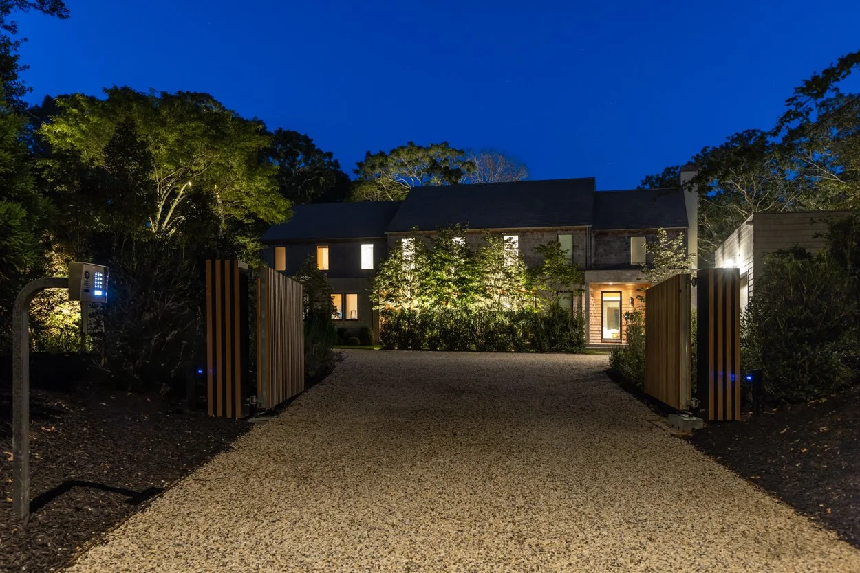 A gravel driveway leading to a modern house at night, surrounded by lush trees and bushes, with exterior lighting illuminating the entrance and front yard.