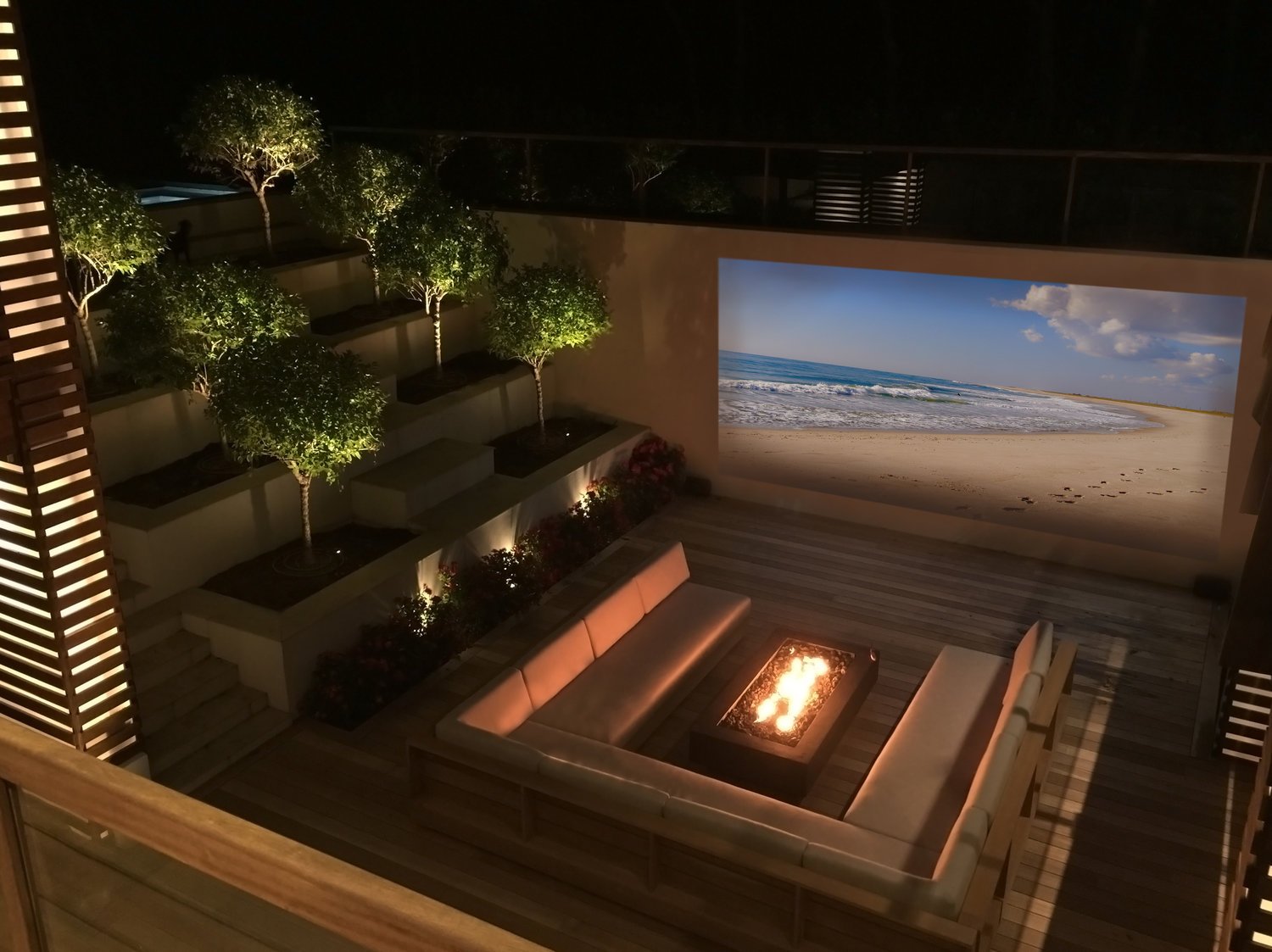 Outdoor patio with a fire pit, beige sectional sofa, and a large wall projection of a beach scene with sand and ocean waves at night.