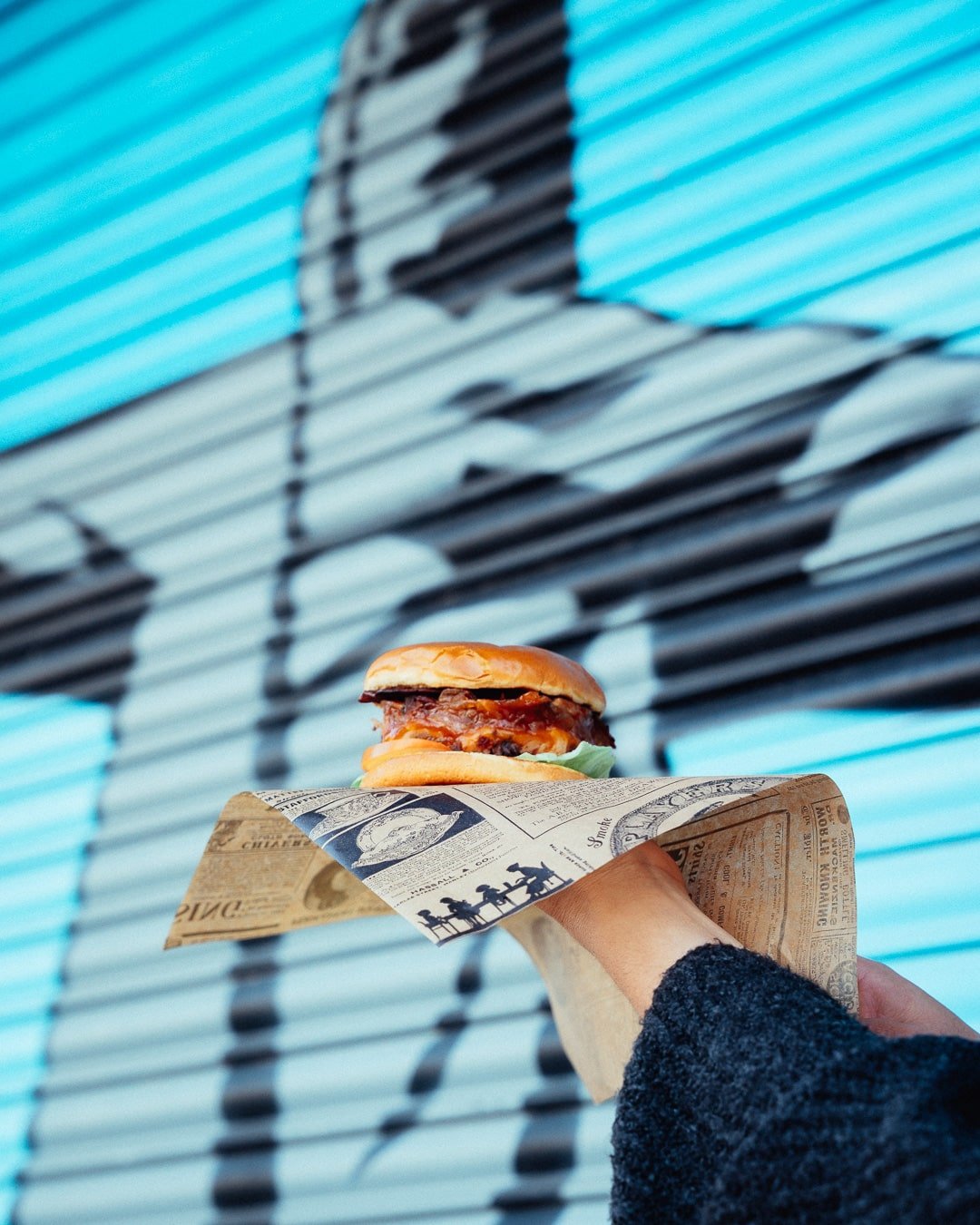 Una mano sostiene una hamburguesa envuelta en papel impreso, con un mural azul y negro de fondo.