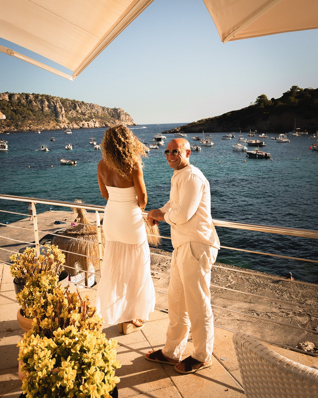 Vistas panorámicas desde la terraza del Restaurante Es Raor en Sant Elm, Andratx, Mallorca. Una pareja, vestida de blanco, admira la bahía llena de barcos fondeados al atardecer.