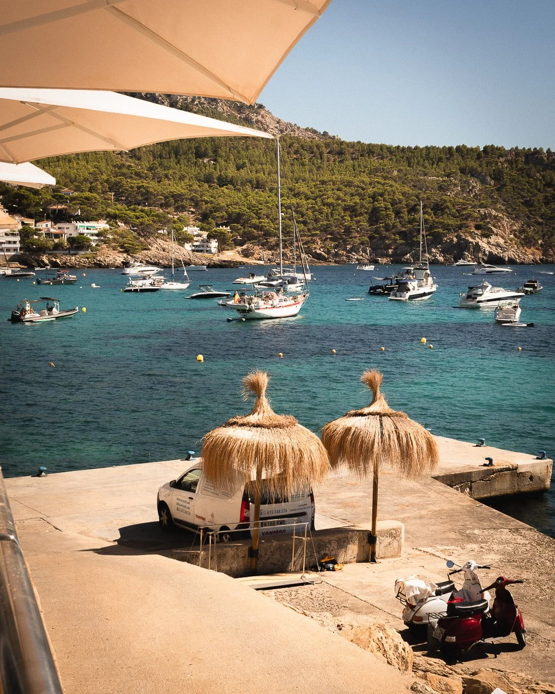 Vistas panorámicas de la cala de Sant Elm con aguas turquesas, barcos fondeados y el muelle tradicional, vistas desde la terraza del Restaurante Es Raor en Andratx.