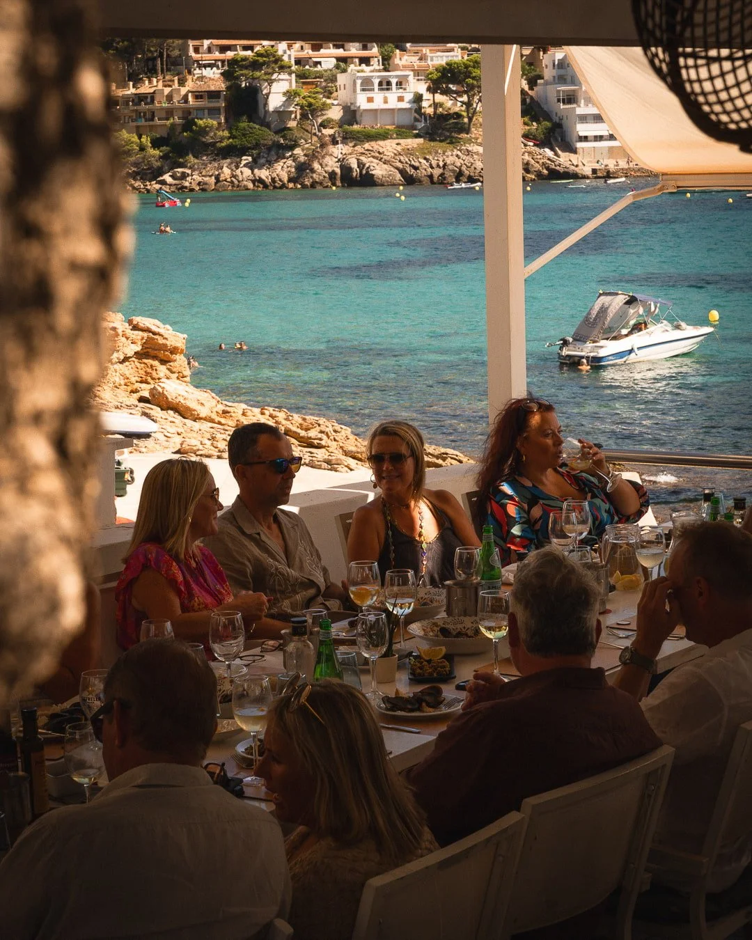Grupo de comensales disfrutando de una comida y vino en la terraza del Restaurante Es Raor, con vistas a las aguas turquesas de Sant Elm, Andratx.