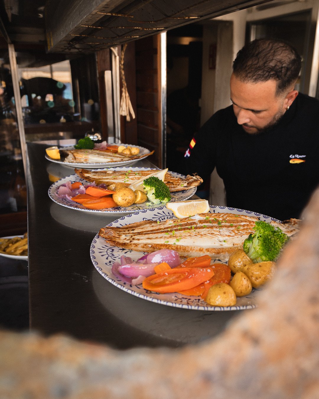 Chef del Restaurante Es Raor en Sant Elm revisando tres platos de pescado fresco a la plancha con guarnición de verduras y patatas asadas en el mostrador de la cocina.