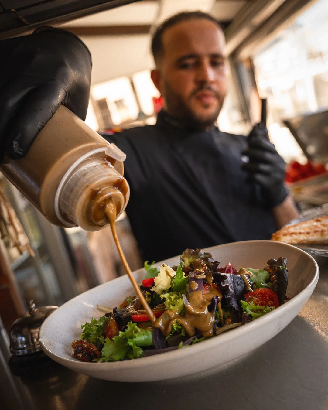 Chef del Restaurante Es Raor en Sant Elm, vistiendo uniforme y guantes negros, vertiendo un aderezo artesanal sobre una ensalada fresca de brotes verdes y tomate.