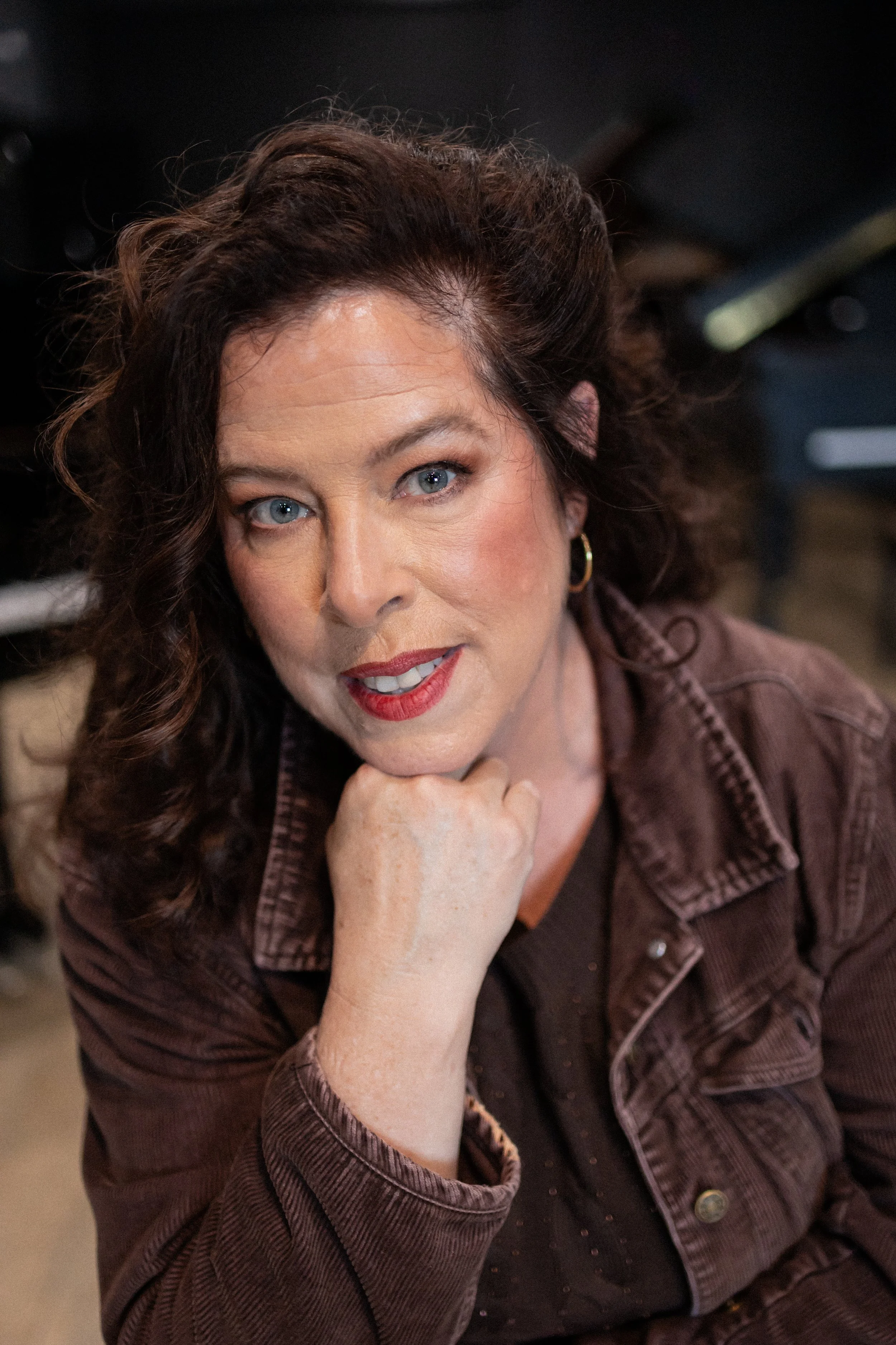 A woman with curly brown hair and blue eyes wearing a brown jacket and red lipstick, resting her chin on her hand and looking at the camera.