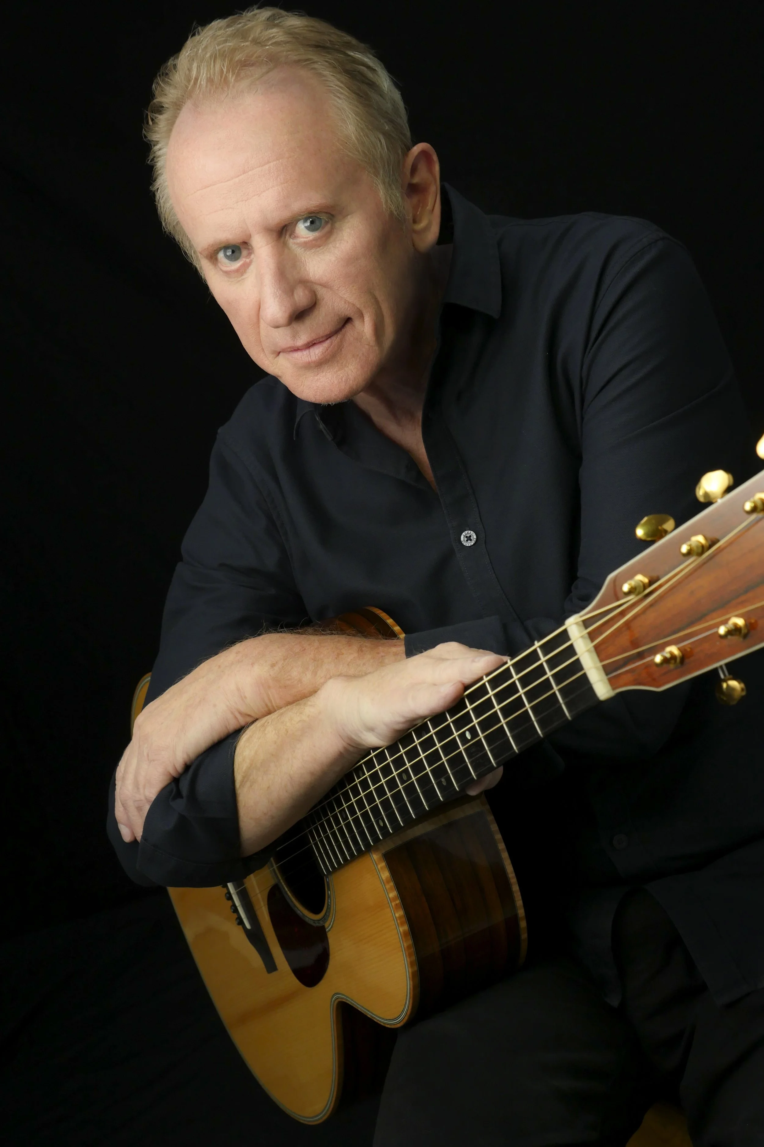 A man with light skin, blonde hair, and blue eyes holding an acoustic guitar against a black background.