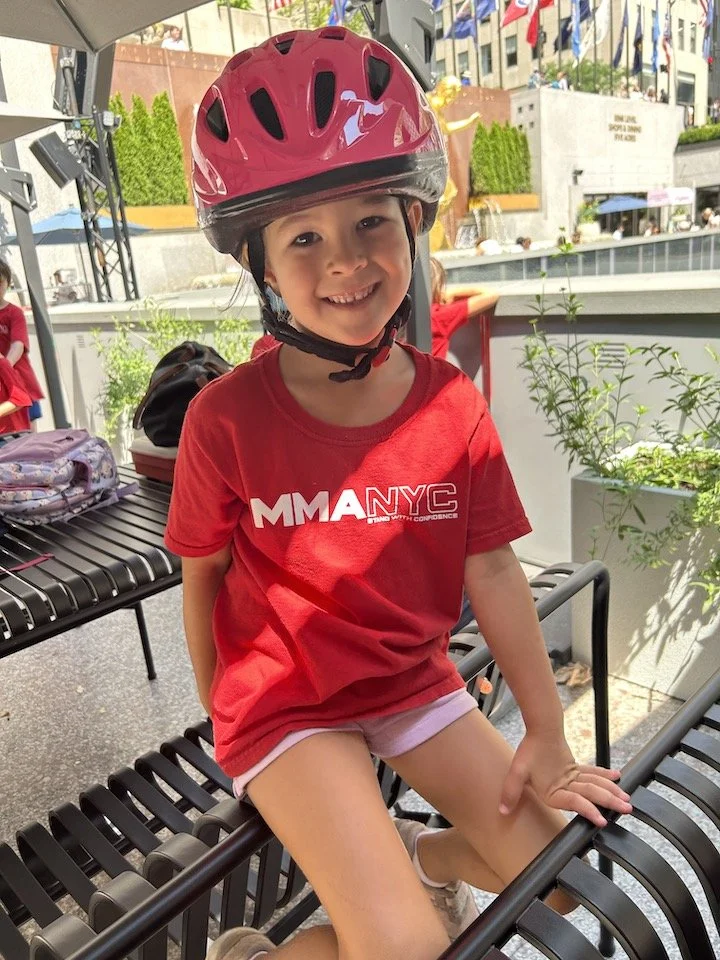 Child wearing a red helmet and t-shirt sitting on a bench, outdoors with flags in the background.