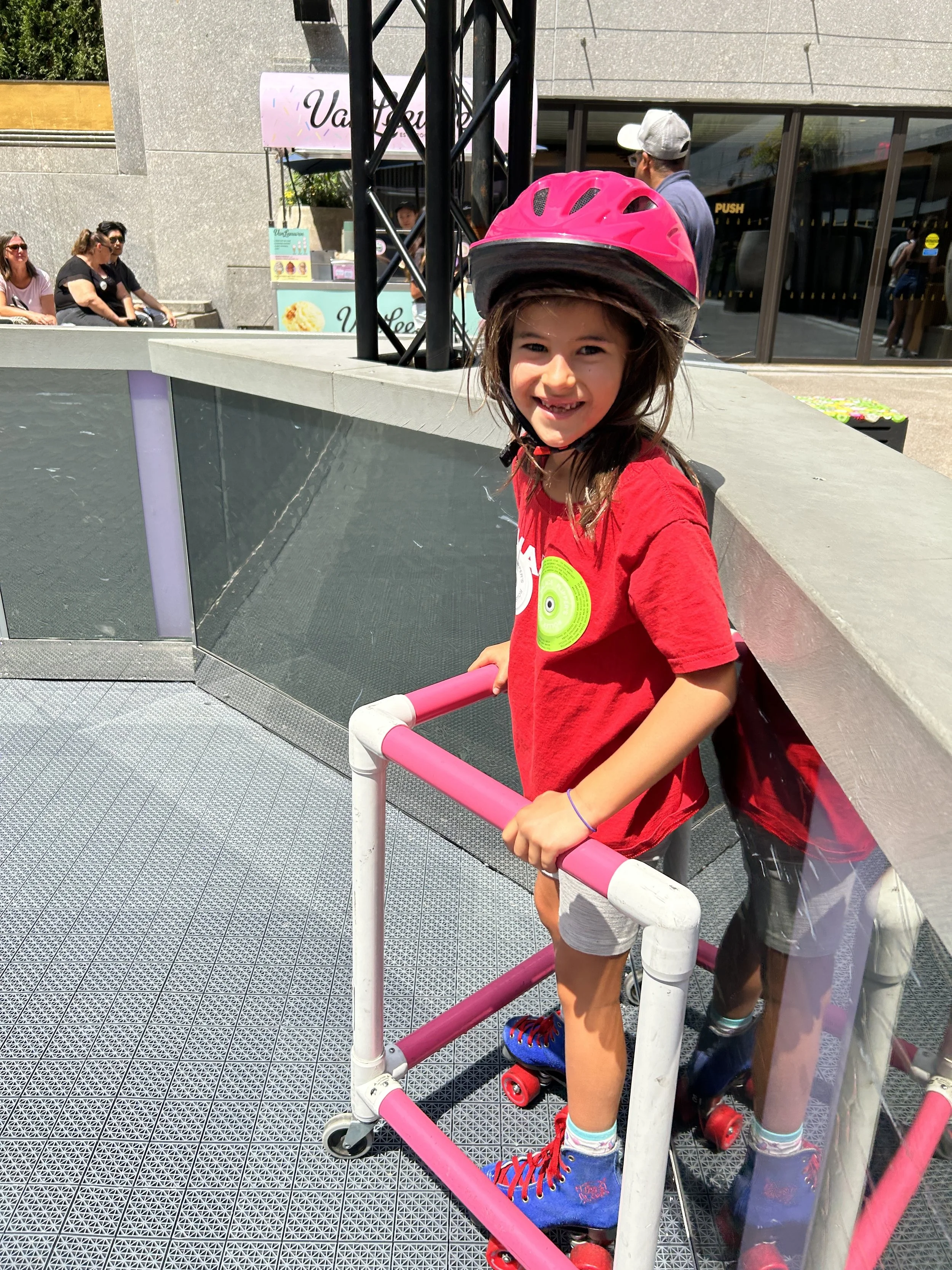 Child roller skating with pink helmet and skate aid on outdoor rink at Rockefeller Center