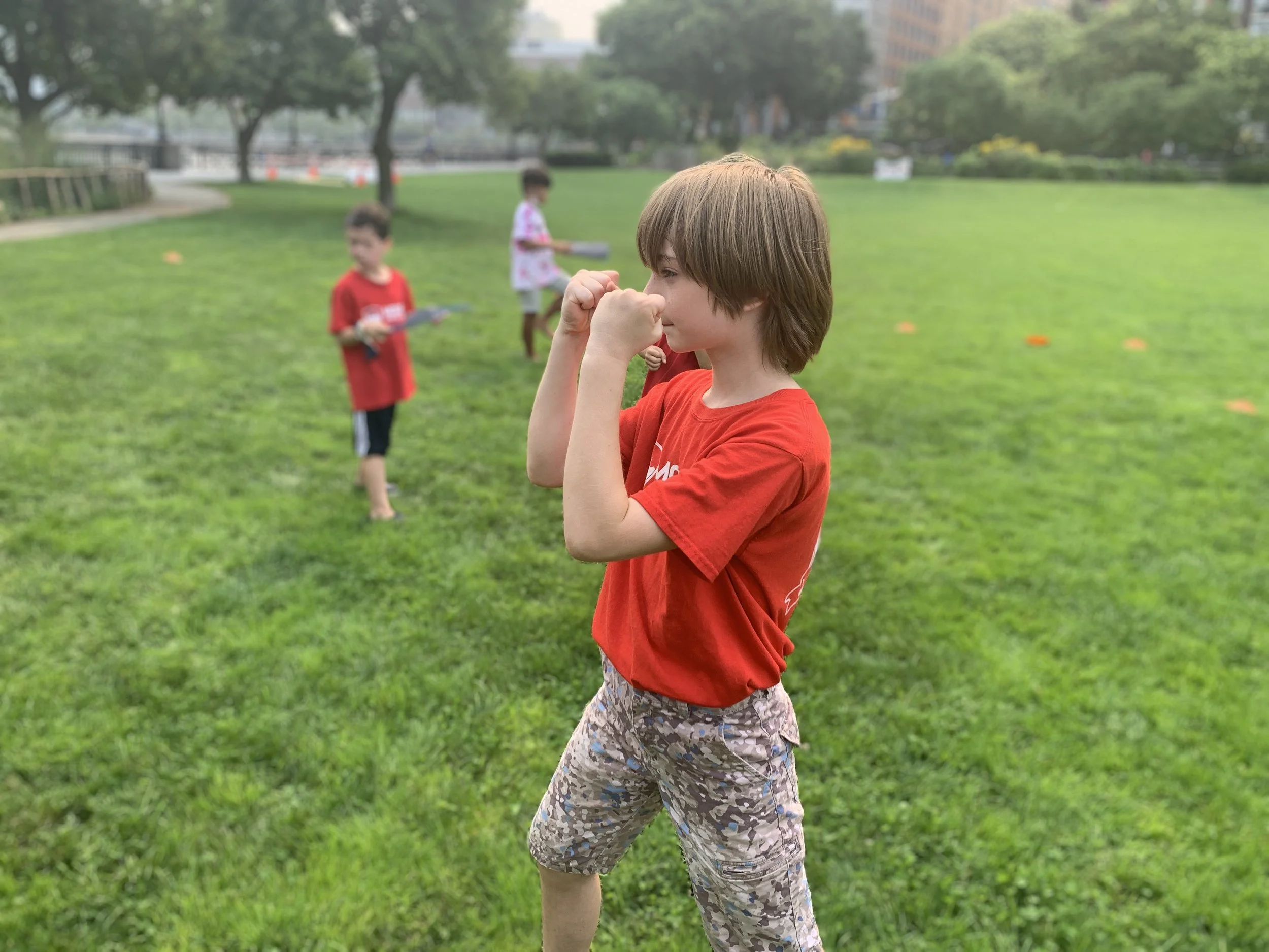 Children playing on a grassy field, one boy in foreground with red shirt and patterned shorts, others in background.