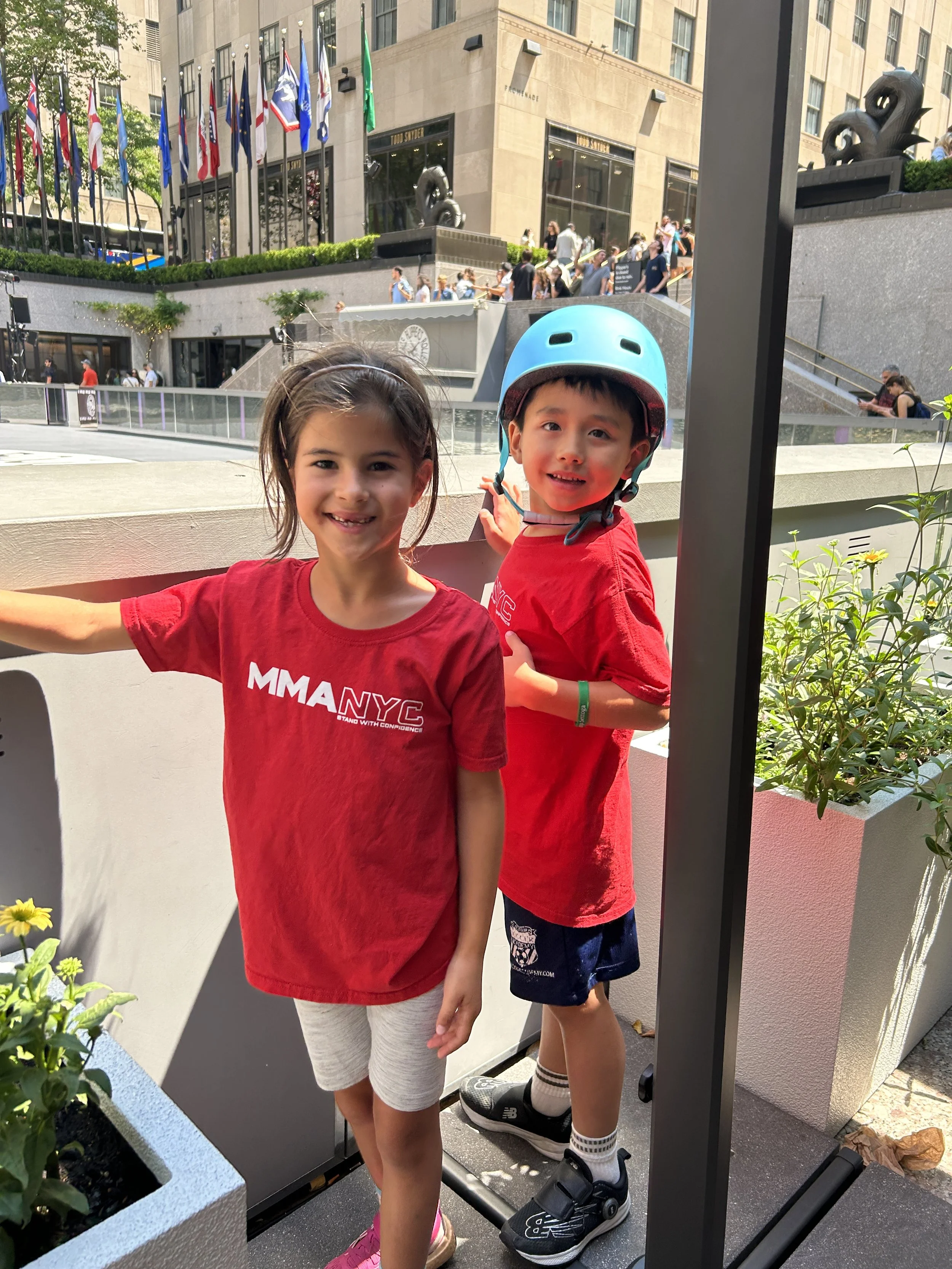 Two children in red shirts, with one wearing a blue helmet, pose and smile at the camera outdoors. Behind them is a plaza area with flags at Rockefeller Center