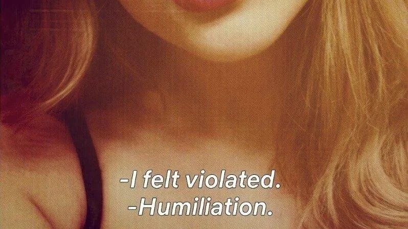 Close-up of a woman with wavy red hair and a black strap on her shoulder, with text overlay reading, 'I felt violated. Humiliation.'