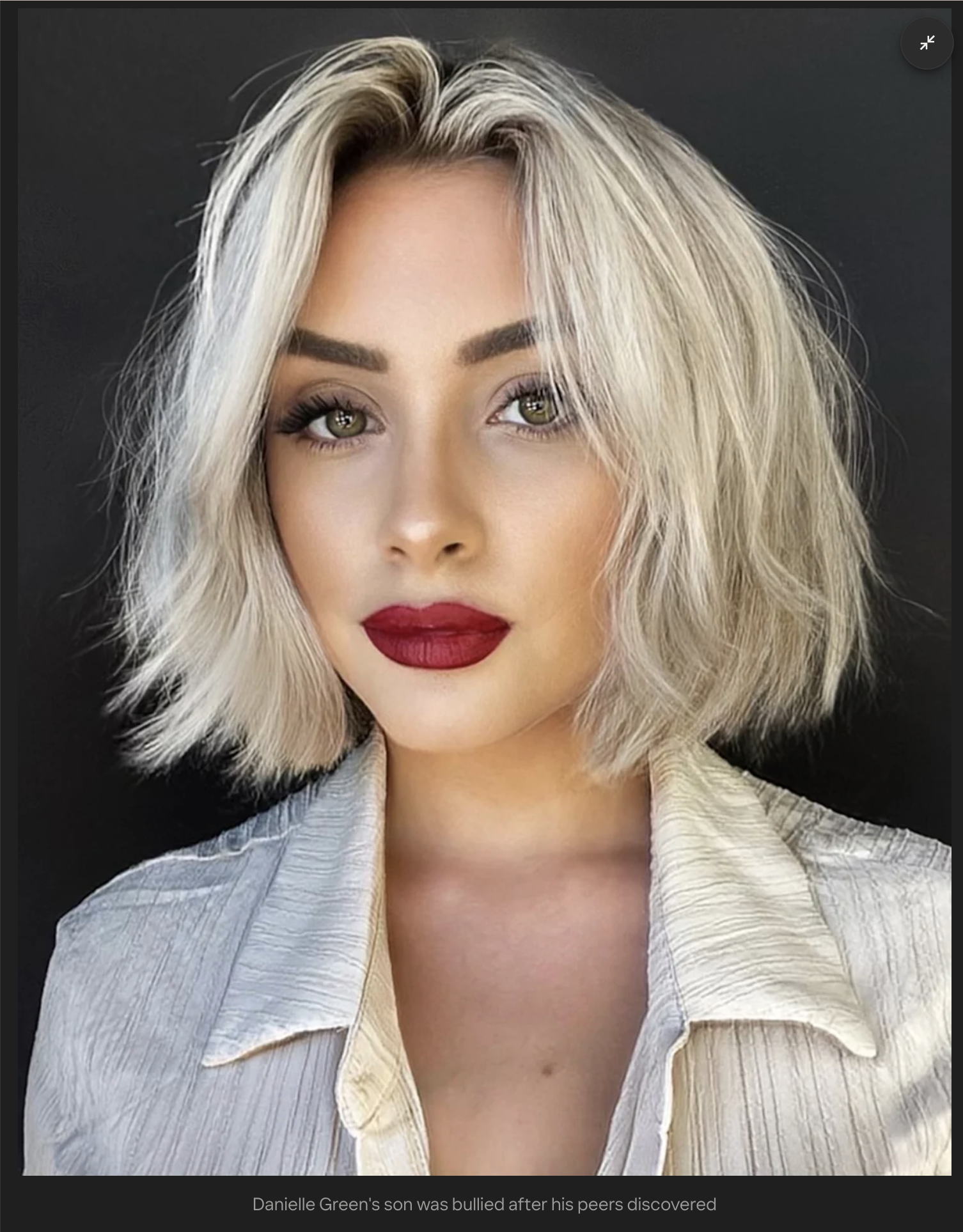Close-up of a woman with blonde wavy hair, wearing a light-colored striped shirt, with makeup including red lipstick and neutral eyeshadow, against a dark background.