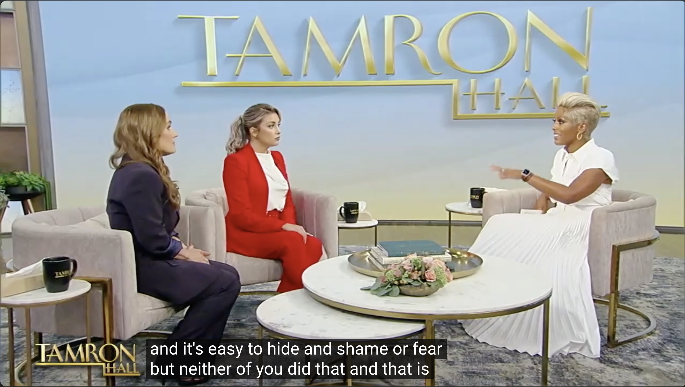Three women seated in a living room, engaged in conversation. The woman on the right has short platinum blonde hair and is smiling, wearing a white outfit. The woman in the middle has wavy gray hair and is wearing a red blazer and pants. The woman on the left has long reddish-brown hair and is dressed in a dark color. There are coffee mugs, a book, and a floral arrangement on a white round coffee table in front of them. The background has a light blue wall with large yellow text that reads "TAMRON HALL."