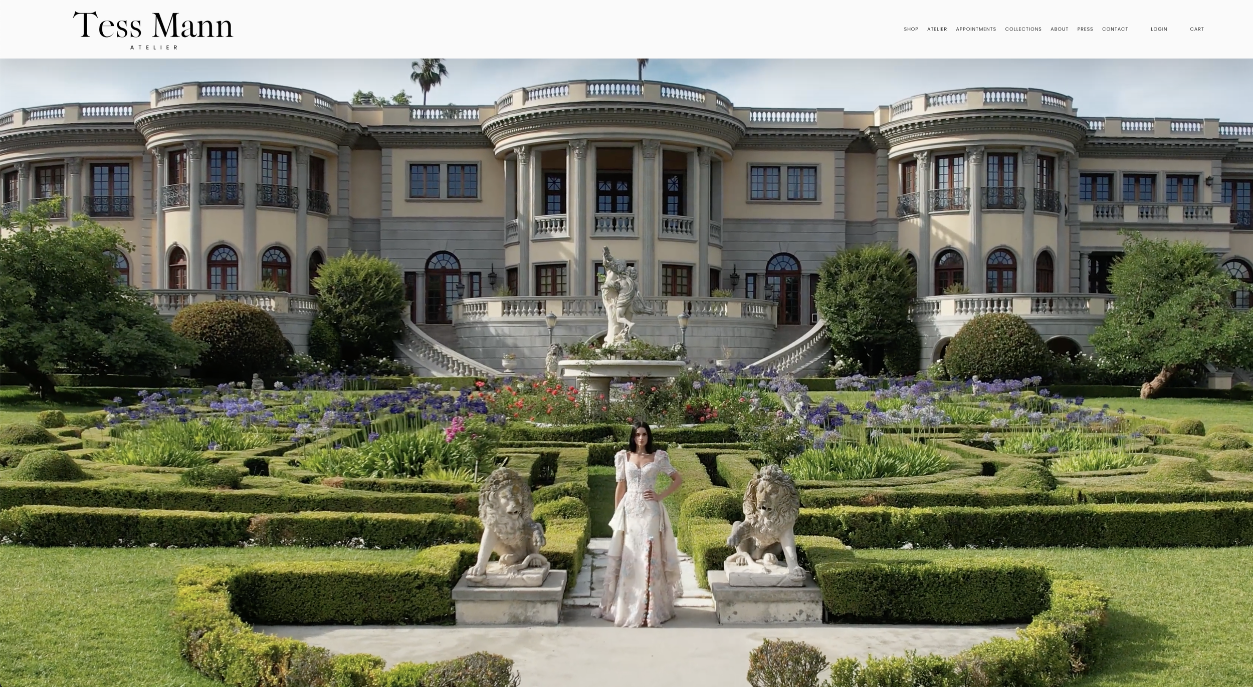 A large mansion with classical architecture and a grand staircase leading up to it. The front yard features a formal garden with neatly trimmed hedges, flowering plants, and classical statues, including lion sculptures and a fountain with a statue of a woman. A woman in a white dress stands among the sculptures in the garden.