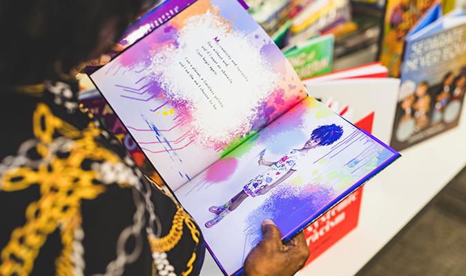 Person holding an open book with colorful splashes of paint, featuring a young girl on one page, in a bookstore or library.