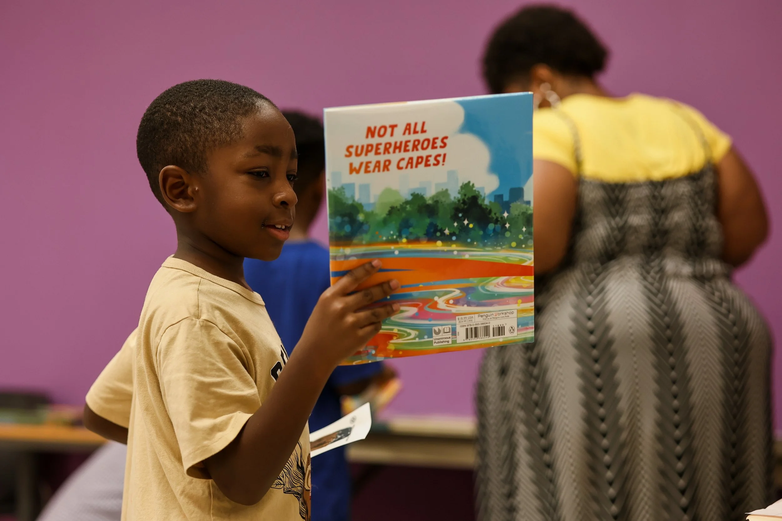 A young girl with short hair holding a colorful book titled 'Not All Superheroes Wear Capes!' in a room with purple walls, with other children in the background.