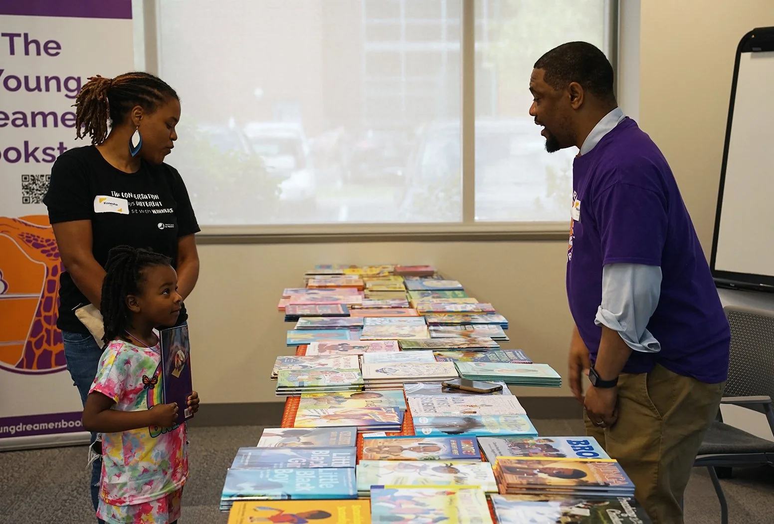 A woman and a young girl are talking to a man at a table filled with colorful books. The woman has dreadlocks and is wearing a black T-shirt, the girl is holding a book, and the man is wearing a purple T-shirt. They are inside a room with a large window and a banner in the background that reads 'The Young Dreamers Bookstore'.