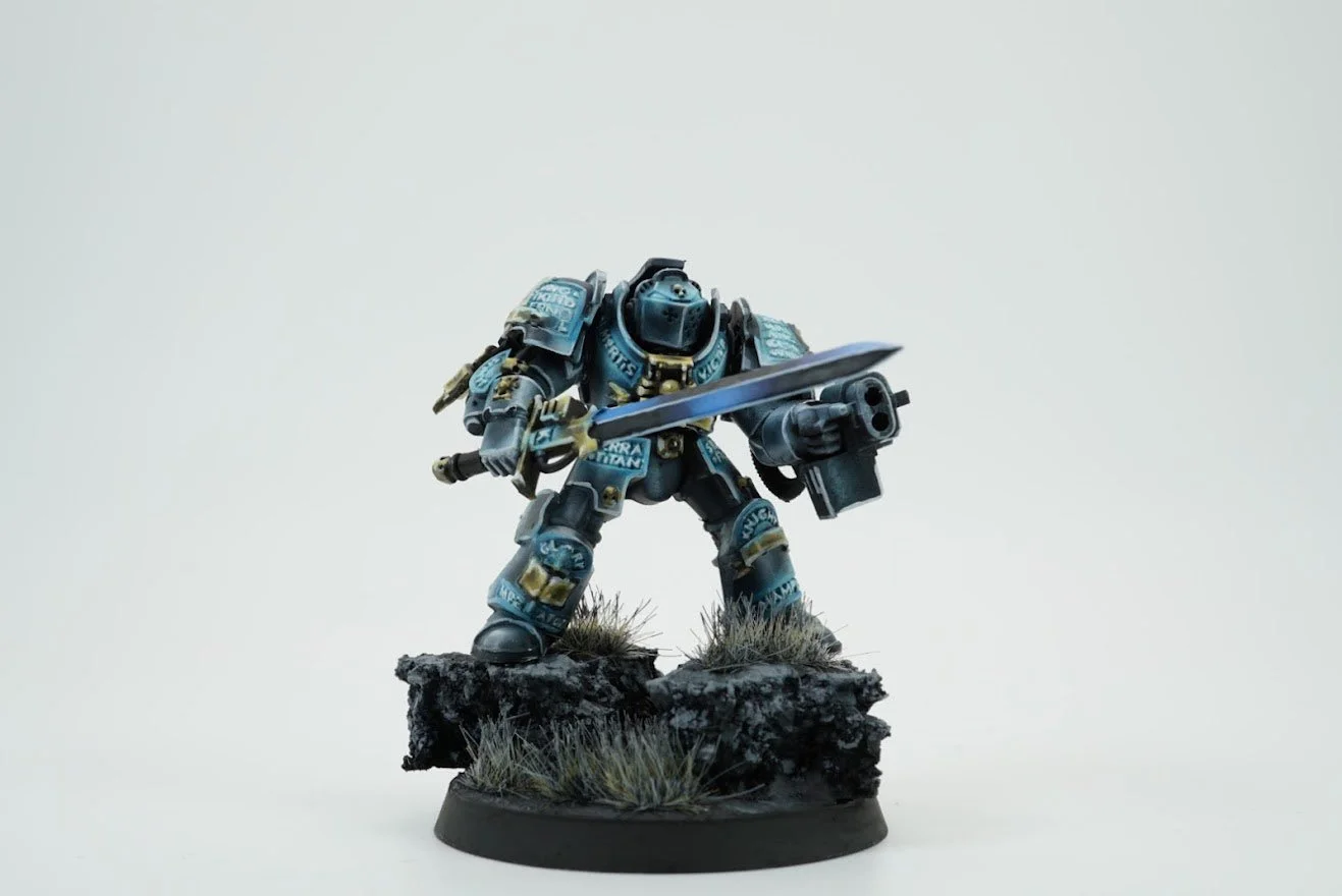 Miniature model of a space marine soldier in futuristic armor, holding a sword and gun, standing on a rocky base with grass details.