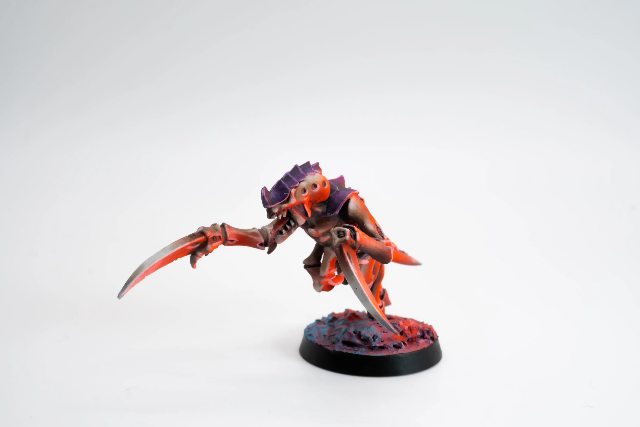 A painted miniature figurine of a sci-fi alien creature with a crab-like body, multiple limbs, and a purple crest head, standing on a textured base with red and purple colors.