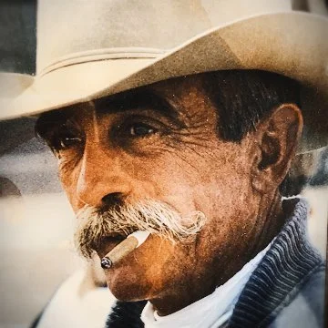 Close-up of an older man with a mustache smoking a cigarette, wearing a beige cowboy hat and a striped jacket.