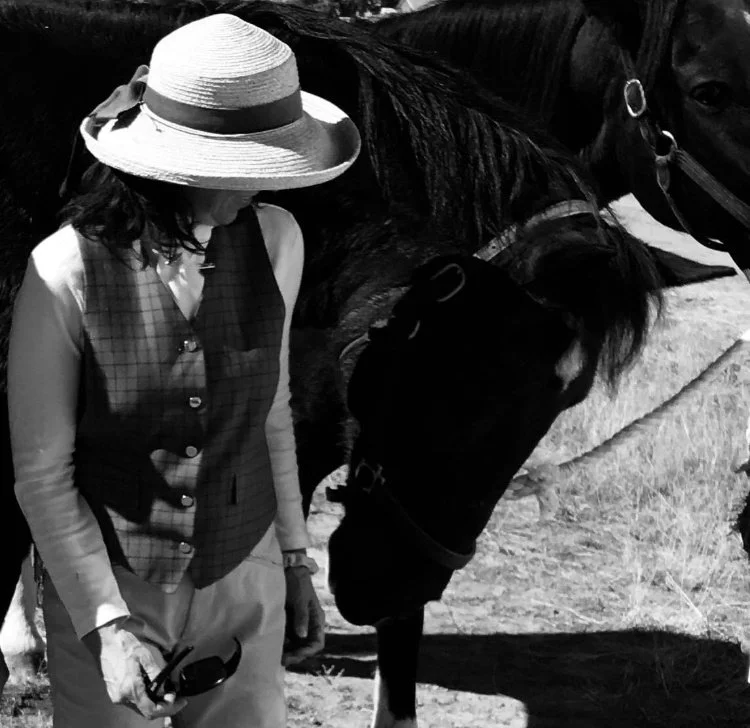 A woman wearing a wide-brimmed hat and plaid vest petting a black horse outdoors.