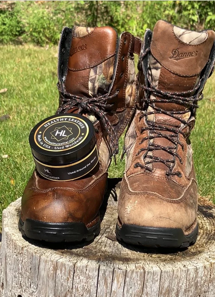 Pair of worn brown leather and camouflage work boots outdoors on a tree stump, with a tin of leather conditioner in front highlighting the before and after application of the Healthy Leather product.
