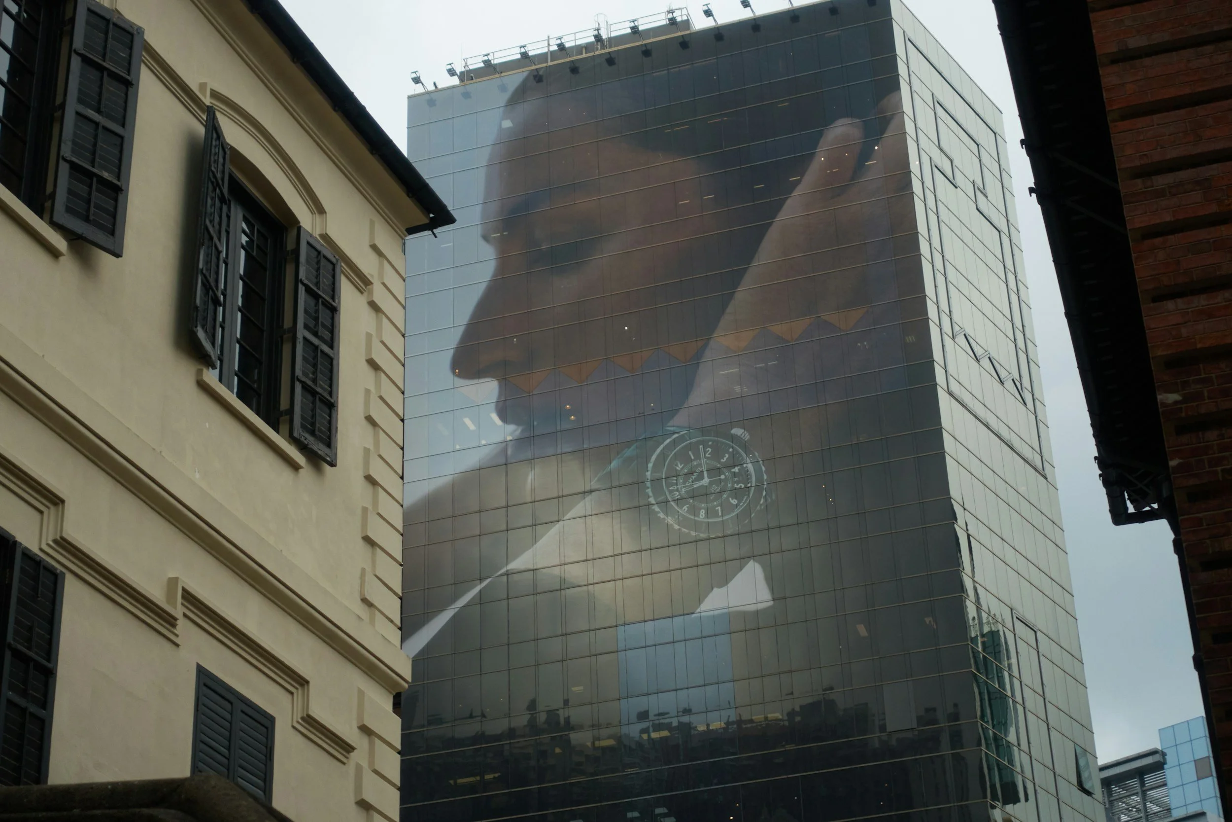 A glass skyscraper with reflective windows showing the reflections of an airplane and the sky, along with a giant advert for a watch brand. The building is surrounded by older buildings with classic architecture and black shutters on the windows.