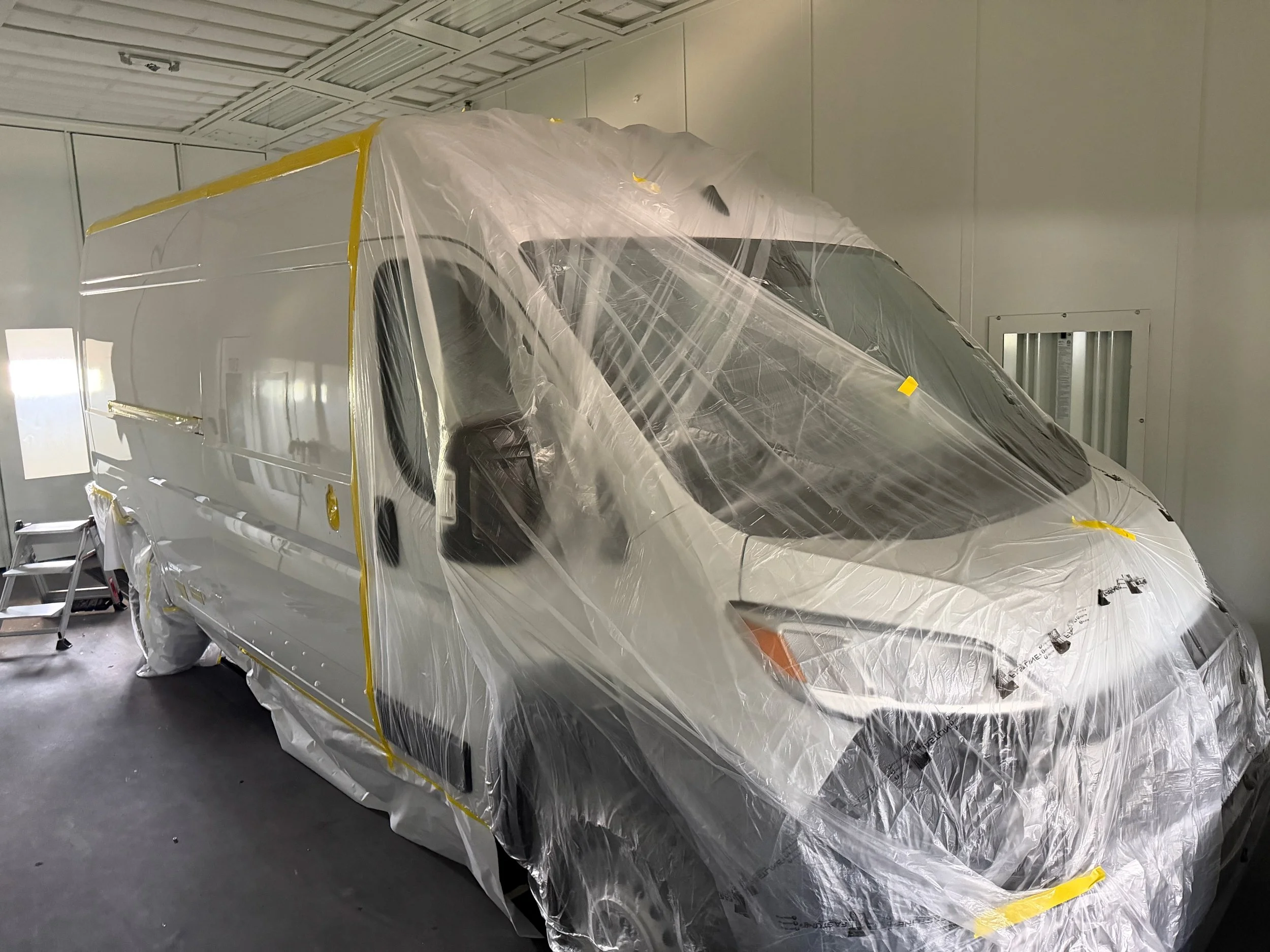 A white van inside an industrial paint booth, covered in clear plastic sheeting with yellow tape securing it, preparing for painting.