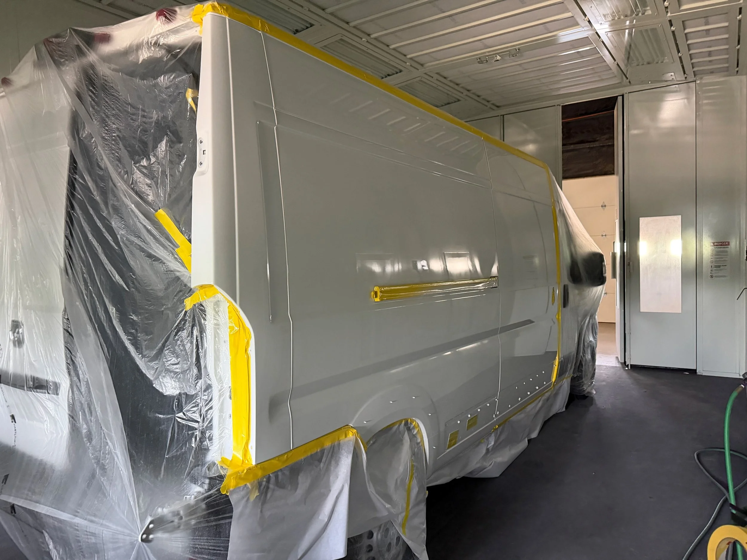 A large van with plastic and yellow tape in an high top paint booth for collision repair refinishing