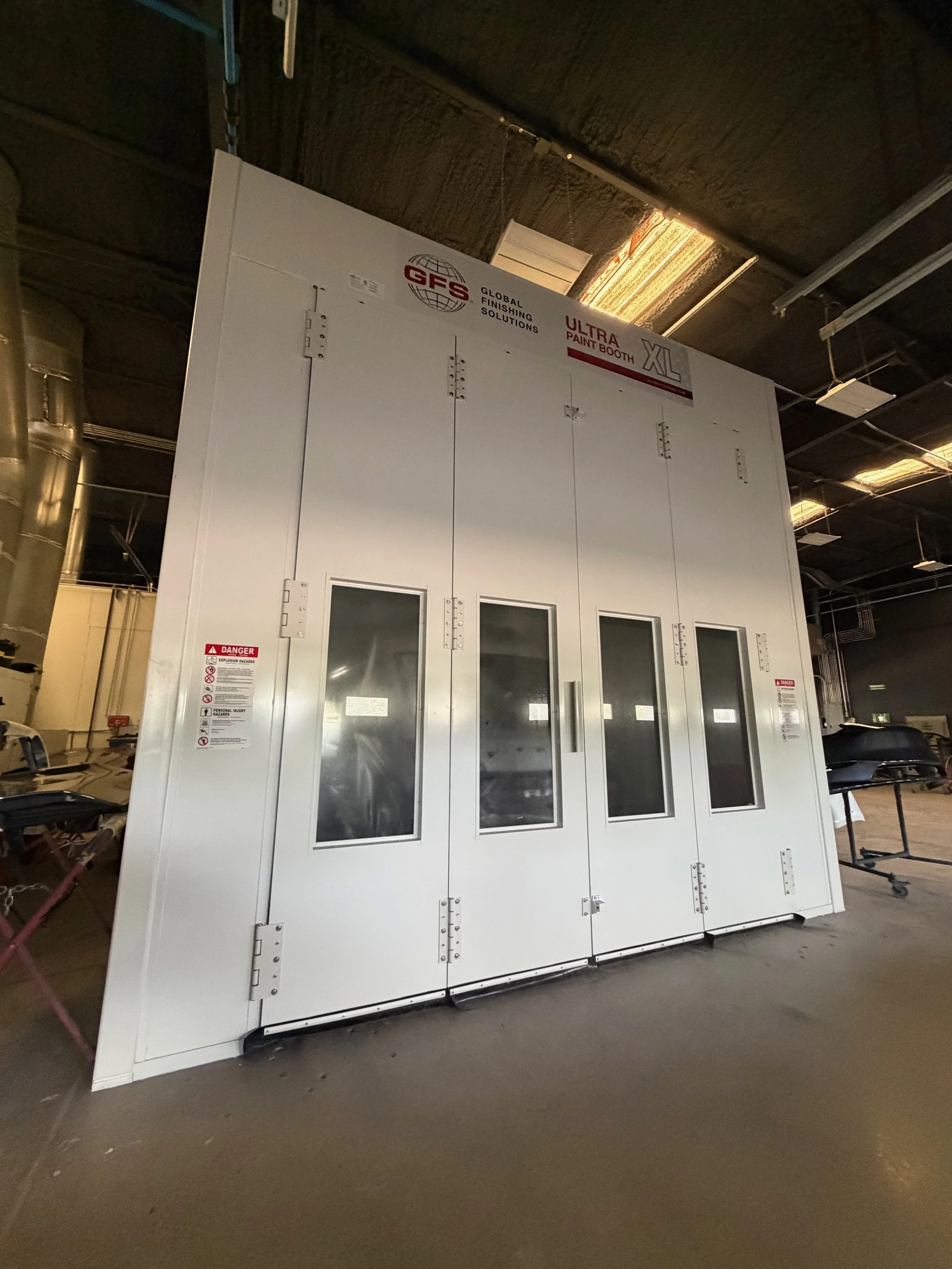 A large white industrial paint booth with glass window panels on doors, warning labels on the side, and a ceiling light, located in a warehouse or workshop setting.