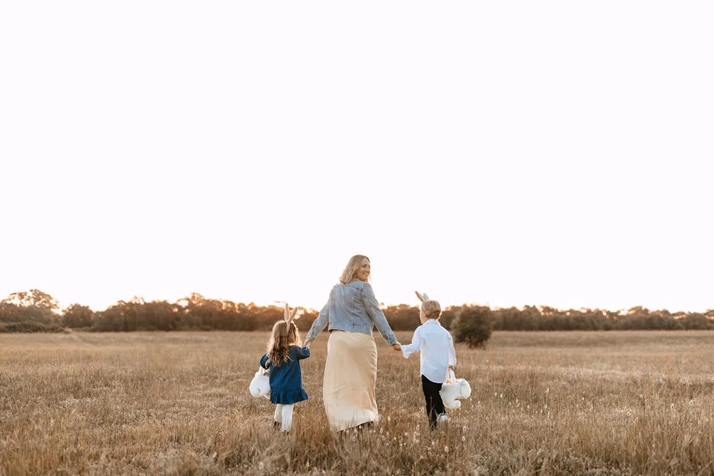 A woman walking with two children in a field at sunset, holding hands, carrying Easter baskets and wearing bunny ears.