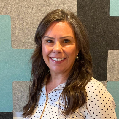 Headshot of a woman with long brown hair wearing a white blouse, smiling, standing in front of a geometric patterned background.