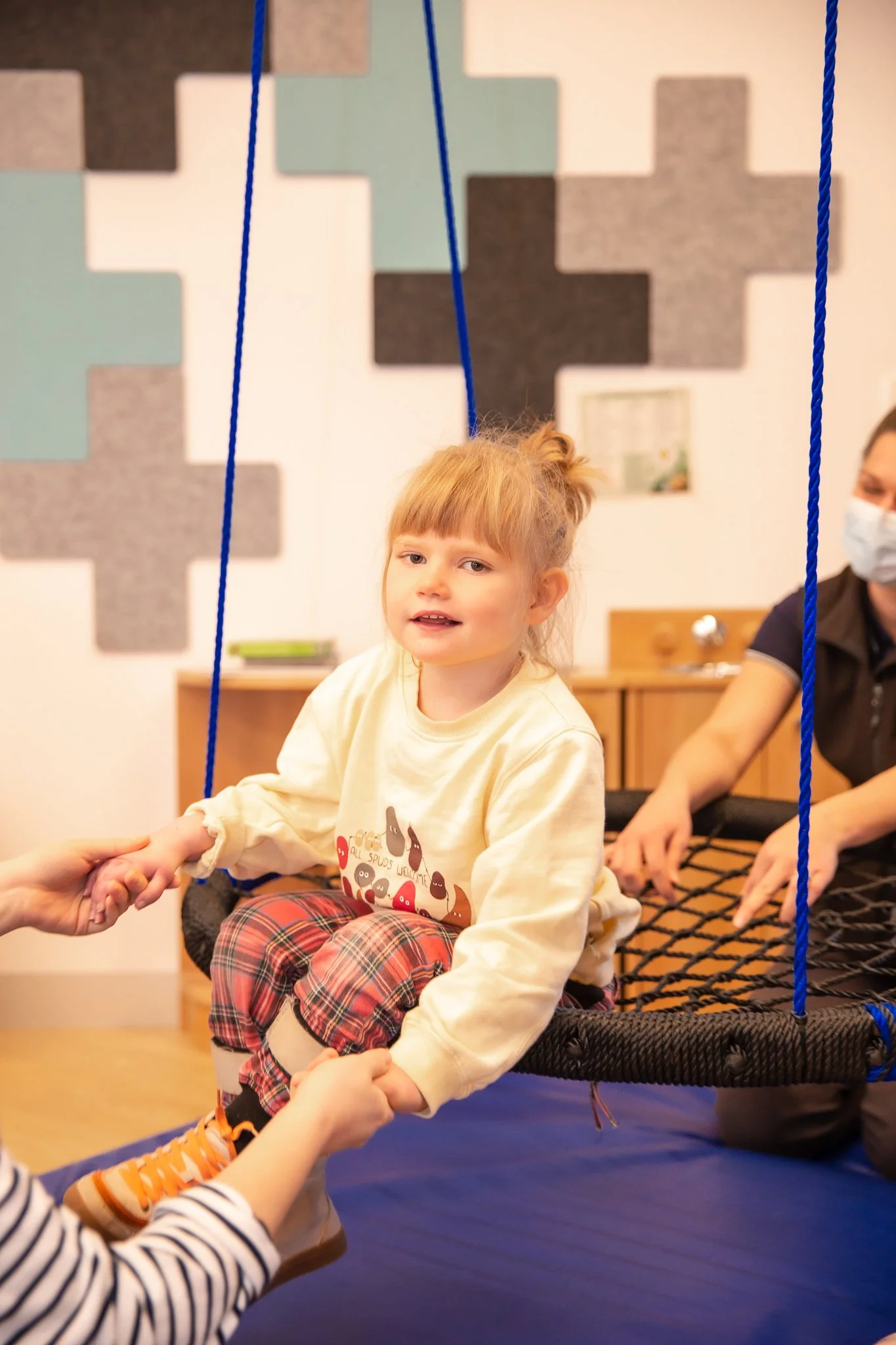 A young girl with red hair and bangs sitting on a black net swing, holding hands with an adult. She is wearing a cream-colored sweatshirt with a graphic design, plaid pants, and orange sneakers. An adult woman in a face mask is assisting her from the side. The background features a wall with geometric patterns in shades of gray, teal, and beige.