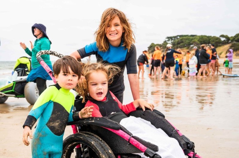 three children, one using a beach wheelchair, smile at the camera while at the beach.