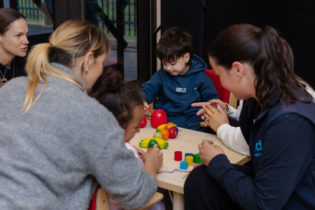 Children and adults sitting around a table playing with colorful toys, engaging in a learning activity indoors.