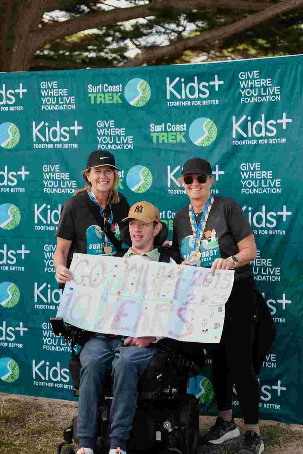 young man using wheelchair is flanked by two women. they are all holding up a sign that says 'GO MUM. 10 YEARS' as they pose in front of a media wall for the Surf Coast Trek event.