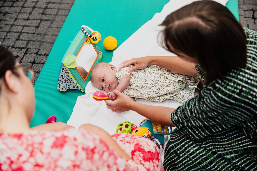 baby lies on its side held on either side by a therapist as it focuses on a red toy rattle