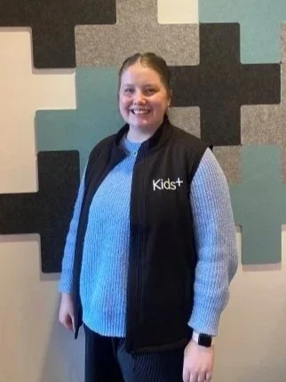 A woman smiling, wearing a black vest with 'Kids+' written on it, standing in front of a colorful, textured wall.
