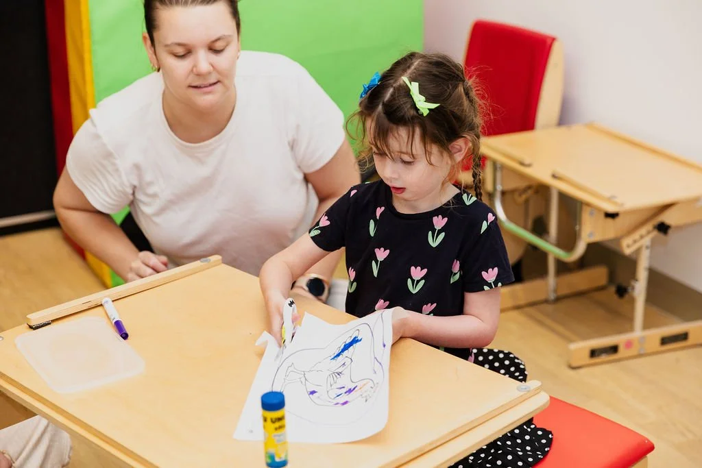 A young girl with pigtails and a woman seated at a table, coloring on a piece of paper with markers in a brightly colored room.