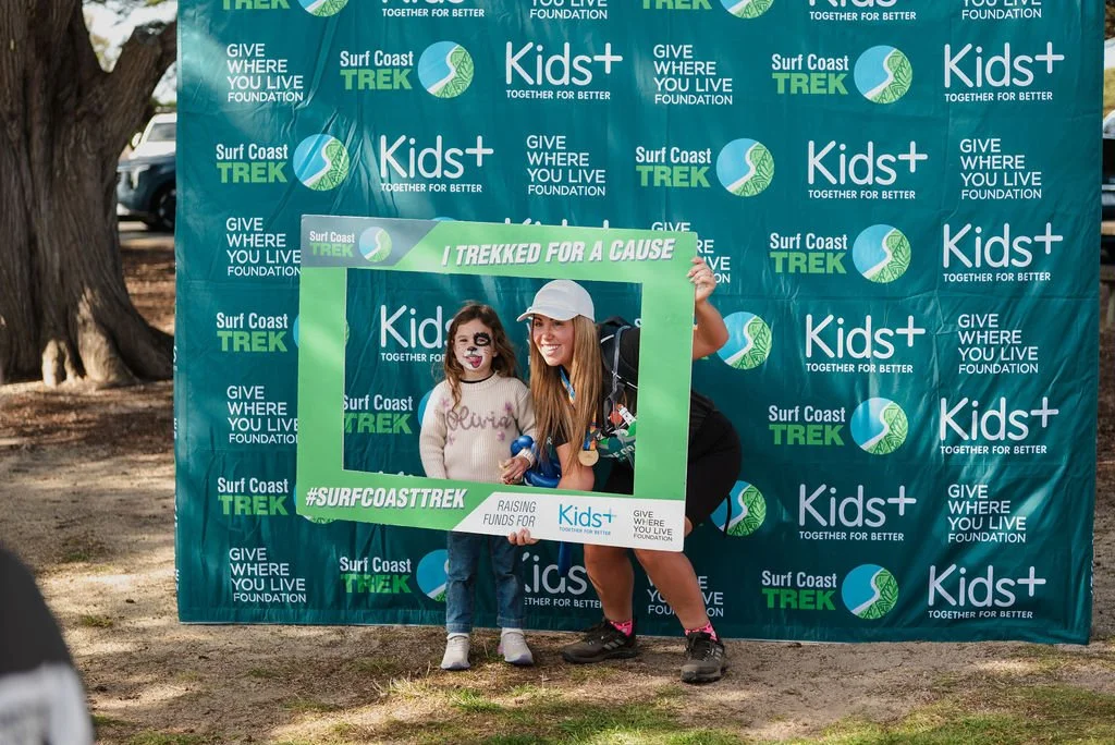 A woman and a young girl are posing behind a large green and white photo frame with the text '#SURFCOASTTREK' at a charity event for Kids+ and Surf Coast Trek. The woman is wearing a white cap and black outfit, while the girl has face paint resembling a dog and is wearing a pink sweater and jeans. They are outdoors with trees and a blue backdrop with multiple event logos in the background.