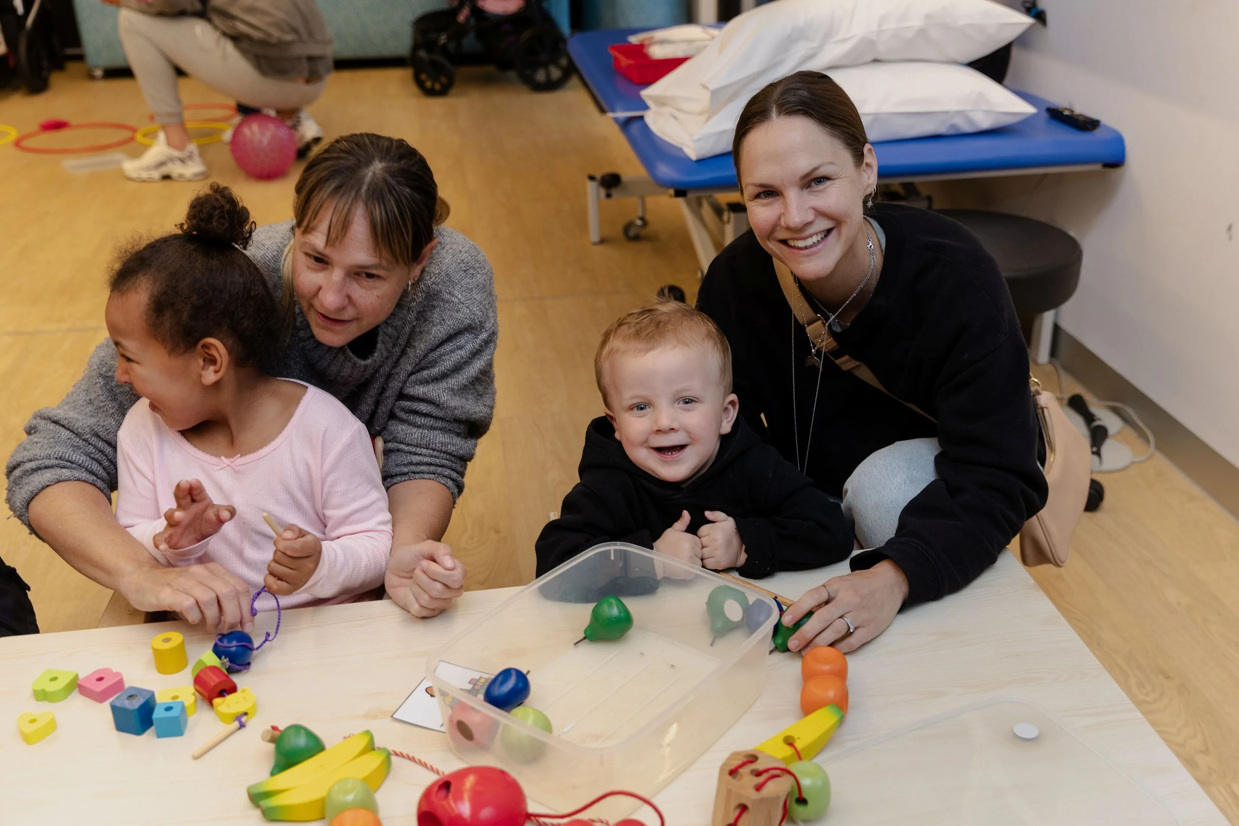 Two children and two women sitting at a table with colorful toys, smiling and interacting, in a room with a bed in the background.